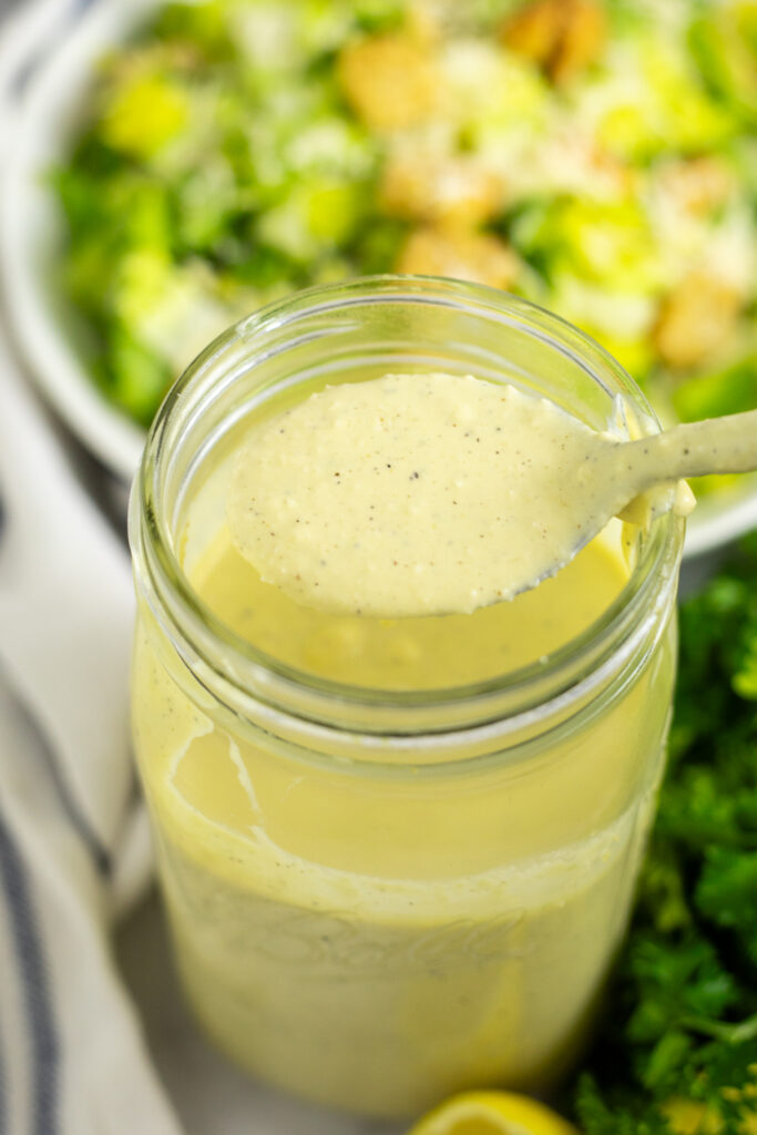A large mason jar filled with homemade caesar dressing and a spoon taking a scoop of it. There is a green salad behind.