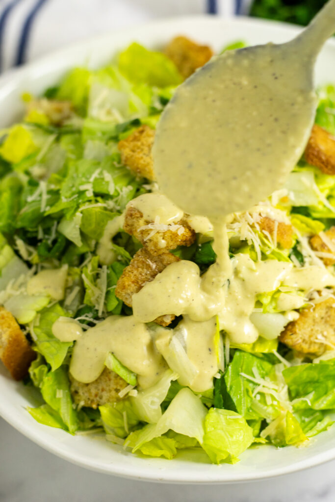 A metal spoon drizzling homemade caesar dressing on top of a green salad with parmesan and croutons.