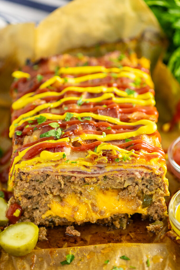 Bacon Cheeseburger Meatloaf with cheese oozing out on a wooden cutting board topped with ketchup and mustard and fresh parsley.