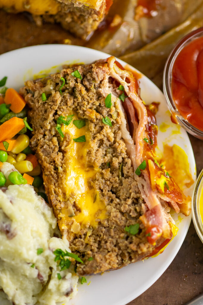 A slice of bacon cheeseburger meatloaf with gooey cheese in the middle on a white plate next to mashed potatoes and mixed vegetables.