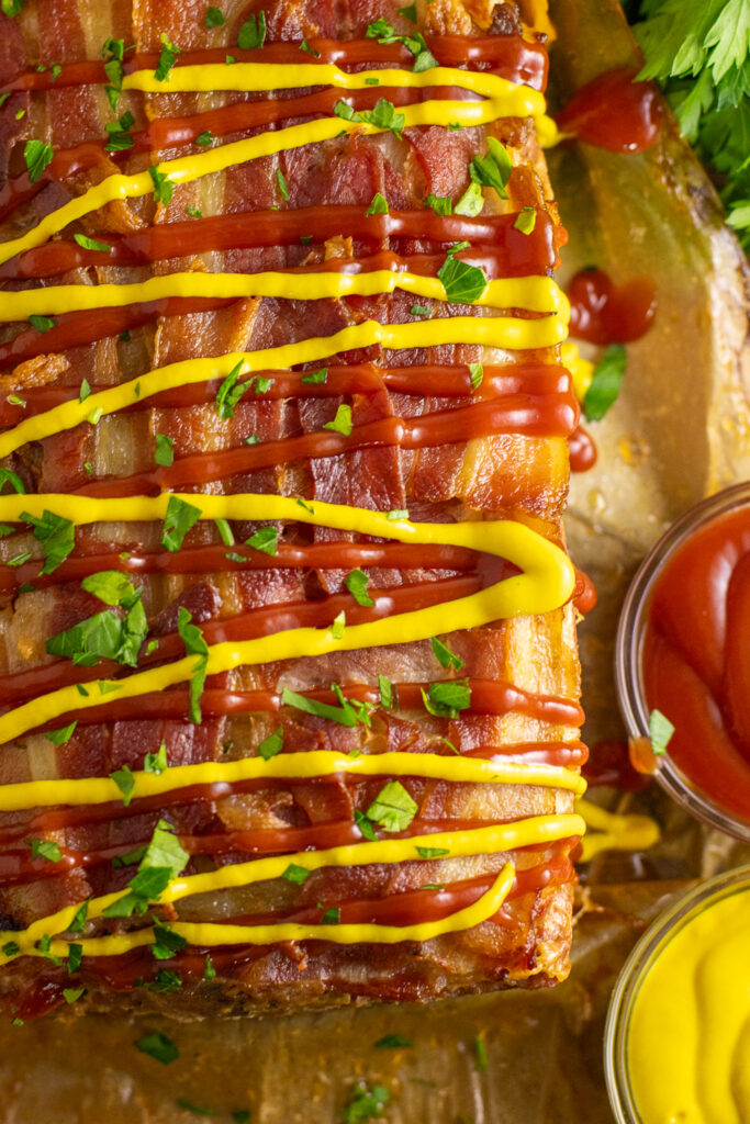 Overhead view of bacon cheeseburger meatloaf topped with bacon and drizzled with ketchup and mustard on a piece of brown parchment paper.
