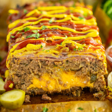 Bacon Cheeseburger Meatloaf with cheese oozing out on a wooden cutting board topped with ketchup and mustard and fresh parsley.
